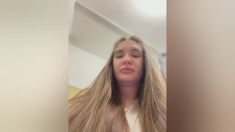 Media: Video of a young Caucasian woman with long, straight blonde hair, wearing a white top, standing in a room with white walls and a ceiling light.