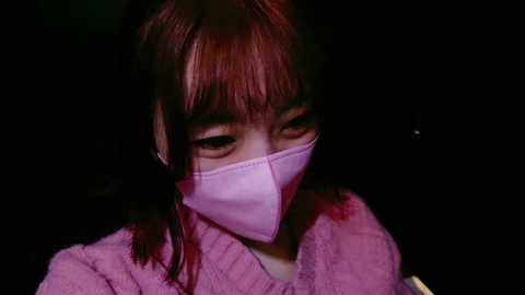 Media: A video of a young Asian woman with shoulder-length dark brown hair, wearing a pink surgical mask and a fuzzy pink robe, set against a dark background.