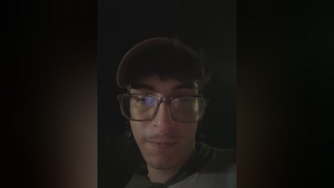 Media: Video of a young man with light skin, wearing glasses, a red cap, and a dark jacket, against a dimly lit background.