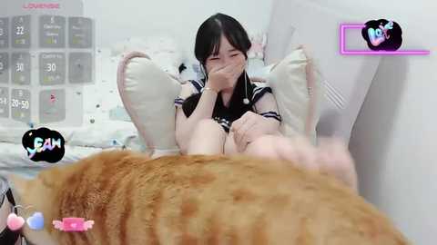 Media: A young Asian woman with long black hair, wearing a black dress, sits on a bed, covering her face with a pillow, while a brown tabby cat approaches her.