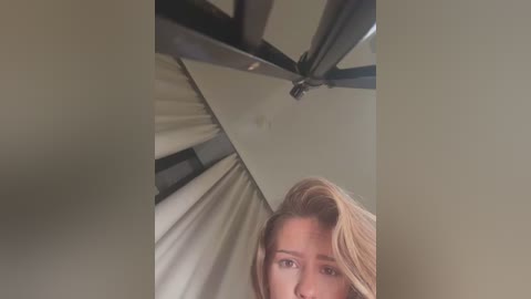 Media: Video of a young woman with fair skin and blonde hair, seen from a low angle, under a white ceiling fan with metallic blades.