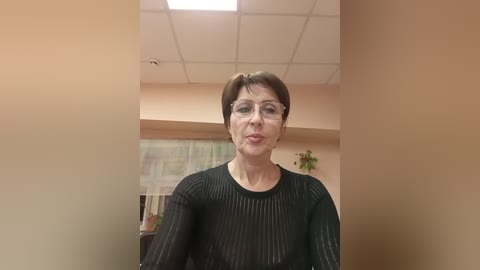 Media: Video of a middle-aged woman with short brown hair, wearing glasses, a black mesh top, and a pensive expression, standing in a beige-walled room with a ceiling light and a framed map on the wall.