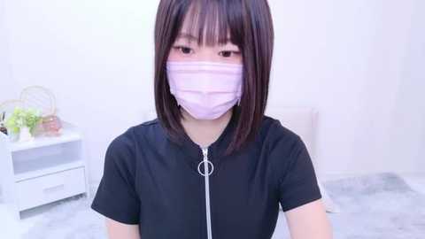 Media: Video of an East Asian woman with shoulder-length, straight brown hair, wearing a black short-sleeved zip-up shirt, light pink face mask, and standing in a minimalist, white room with a white chair and plant.