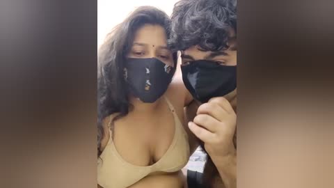 Media: A video of a couple wearing black masks, the woman with long black hair and a beige bra, and the man with curly black hair, both smiling, in a blurred background.