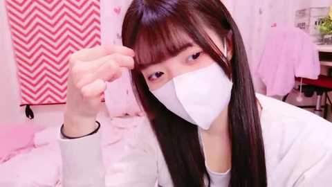 Media: A video of an East Asian woman with long black hair and a white face mask, sitting on a bed with pink and white bedding, adjusting her hair in a pink zigzag patterned room.