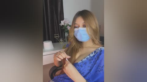 Media: Video of a young woman with light skin, long blonde hair, and blue surgical mask, seated at a desk in a room with a dark curtain, white wall, and a white vase with flowers.