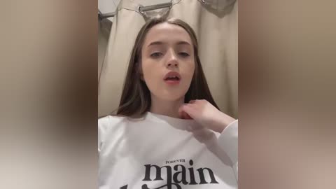 Media: Video of a young, light-skinned woman with long, straight brown hair, wearing a white \"Maine\" T-shirt, sitting in a bathroom with beige shower curtain and towel rack in the background.