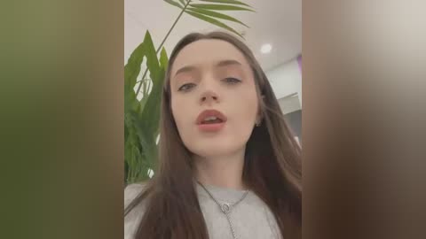 Media: Video of a young woman with long brown hair, fair skin, and light makeup, wearing a gray sweater, looking directly at the camera in an indoor setting with green plants and soft lighting.