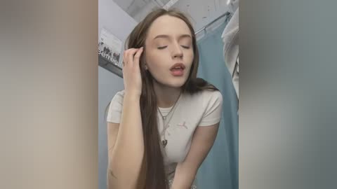 Media: Video of a young Caucasian woman with long brown hair, wearing a white T-shirt, touching her ear in a bathroom with a blue shower curtain.