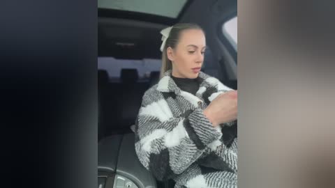 Media: Video of a light-skinned woman with long blonde hair tied back, wearing a black and white checkered blanket, seated in a car, holding a phone, blurred background.