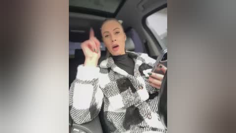 Video of a blonde woman in a black-and-white checkered coat, holding a smartphone, giving a middle finger from the driver's seat of a car.