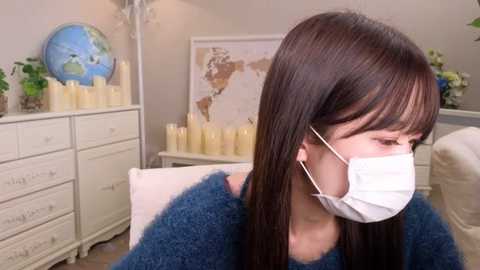 Media: Video of a woman in a blue sweater, wearing a white face mask, seated indoors. Background features a white dresser, candles, and a framed world map.