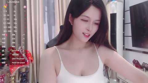 Media: A video of an East Asian woman with long black hair, fair skin, and small breasts, wearing a white spaghetti-strap top, smiling in a cozy, well-lit room.