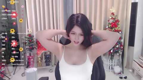 Media: Video of a young Asian woman with short black hair, medium build, wearing a white tank top and black jacket, seated with hands behind head, in a festive room with a decorated Christmas tree and various ornaments.