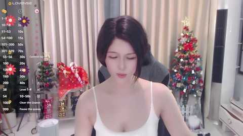 Media: Video of a young East Asian woman with short black hair, wearing a white tank top, sitting in front of a Christmas tree and festive decor, captured in a livestream.