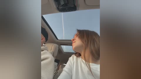 Media: Video of a young couple sitting in a car, with a clear blue sky visible through the sunroof. The woman has long brown hair and wears a white top, while the man, with short dark hair, wears a gray sweater.