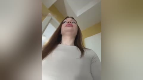 Media: A video of a young Caucasian woman with long brown hair, wearing a white long-sleeve shirt, standing in a room with beige and white walls.