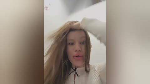 Media: Video of a young woman with fair skin and long, straight blonde hair, wearing a white ribbed sweater. She looks surprised with her hand in her hair, standing indoors in a blurred, dimly lit room.