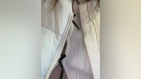 Media: Video of a person's torso in a white, ribbed knit sweater with a black zipper partially open, revealing a light pink shirt underneath. The background is plain, off-white wall.