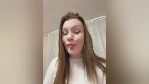 Media: Video of a young woman with long, straight brown hair, wearing a white top, licking a lollipop. She's indoors, with a white curtain and beige wall in the background.