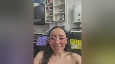 Media: Video of a smiling, topless woman with long dark hair and glasses, standing in a small, cluttered kitchen with white tiled walls, cabinets, and a gas stove.