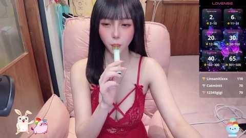 Media: Video of an Asian woman with long black hair in a red lace lingerie, eating a popsicle in a pink chair, with a virtual camera overlay.