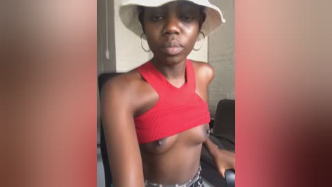 Media: Video of a young, dark-skinned Black woman with small breasts, wearing a red crop top and white bucket hat, sitting in a dimly lit room with beige walls.