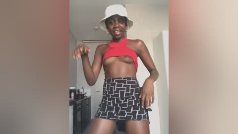 Media: Video of a dark-skinned, slender woman with small breasts, wearing a white bucket hat, red crop top, and black-and-white patterned skirt, in a modern, minimalist room with white walls and a kitchen area in the background.