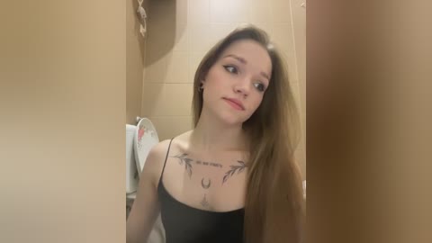 Media: Video of a young Caucasian woman with long, straight brown hair, wearing a black tank top, standing in a beige-tiled bathroom with a crescent moon tattoo on her chest.