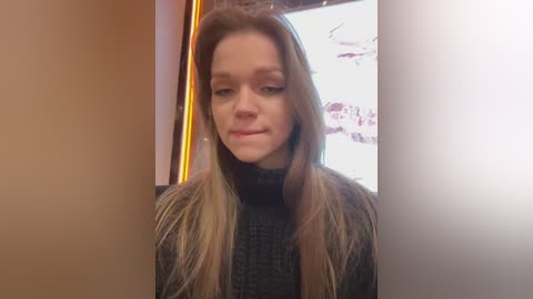 Media: Video of a young Caucasian woman with long, straight, light brown hair, wearing a black turtleneck sweater. She has a neutral expression and is standing in a dimly lit room with beige walls.