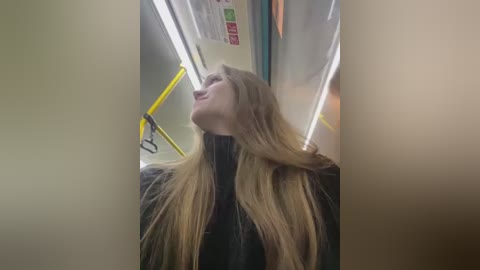 A video of a blonde woman with long, straight hair, wearing a black jacket, looking upwards in a subway car with fluorescent lights and yellow handrails.