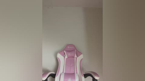 Media: A video of a pink and white gaming chair with the brand name \"Razer\" on the backrest, positioned against a plain, beige wall in a minimalist room. The chair's ergonomic design features adjustable armrests and a cushioned seat.