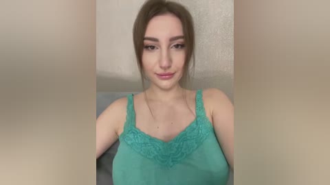 Media: Video of a young Caucasian woman with fair skin and straight, light brown hair. She wears a turquoise lace-trimmed tank top and sits against a beige wall.