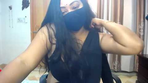 Media: A video of a South Asian woman with long dark hair, wearing a black face mask, black top, and gold bangles. She's adjusting her mask in a simple, cluttered bedroom with wooden furniture and beige curtains.