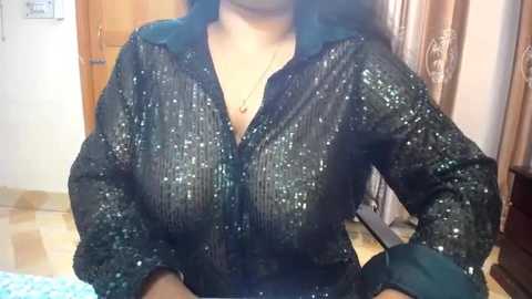 Media: Video of a middle-aged woman with medium skin tone, wearing a shimmering black sequin blouse, revealing cleavage, in a cluttered, dimly lit room with wooden doors and beige curtains in the background.