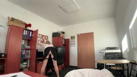 Media: A video of a sparsely furnished office with a person in a black dress bending over a desk, surrounded by bookshelves, a printer, and a computer monitor.