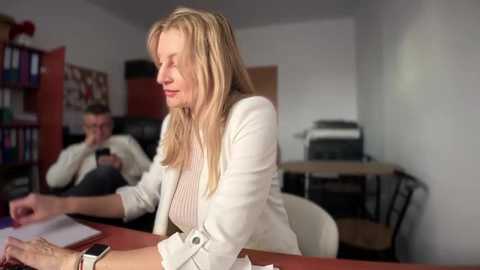 Media: Video of a blonde woman in a white blazer and pink shirt, sitting at a desk, deep in thought. Behind her, a man sits reading a book.