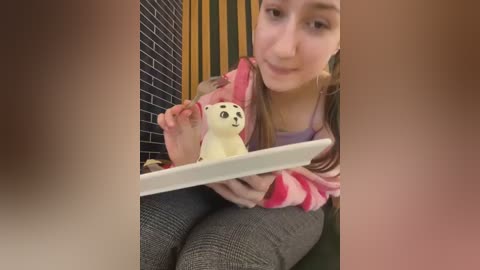 Media: Video of a young woman with light skin, brown hair, and a pink and white striped sweater, sitting on a gray couch. She's holding a tablet and a plush toy bear. Background features a dark brick wall and vertical wooden stripes.