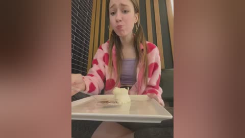 Media: Video of a young Caucasian woman with light skin and long brown hair, wearing a pink heart-patterned robe over a purple tank top, sitting at a table in a modern cafe with dark brick walls and vertical wooden panels. She's making a pouty face while holding a small cup.