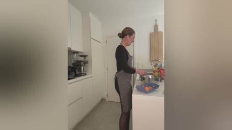 Media: A video of a woman with brown hair in a bun, wearing a black long-sleeve shirt and gray apron, preparing food on a white kitchen counter.