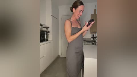 Media: Video of a slim, fair-skinned woman with brown hair in a bun, wearing a gray dress, smiling and looking at her smartphone in a modern, minimalist kitchen with white cabinets and stainless steel appliances.