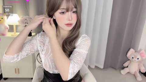 Media: A video of a young Asian woman with fair skin and long brown hair, wearing a white lace blouse, adjusting her hair in a bedroom with gray curtains and a pink plush toy.