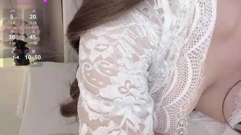 Media: A close-up video of a woman's arm and chest in a white lace robe, with brown hair visible. Background includes a white wall and a small plant.