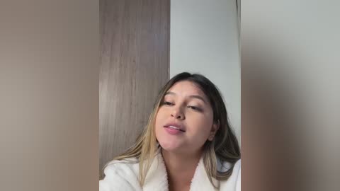 Media: Video of a young woman with light brown hair, wearing a white bathrobe, standing in a beige-tiled bathroom, eyes closed, mouth slightly open, looking relaxed.