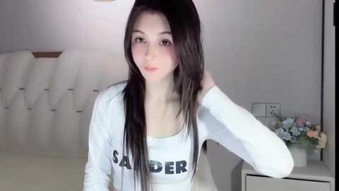 Media: Video of a young Asian woman with long black hair, fair skin, and a slender physique. She wears a white \"SADDER\" T-shirt and sits in a minimalist bedroom with a floral arrangement on a white nightstand.