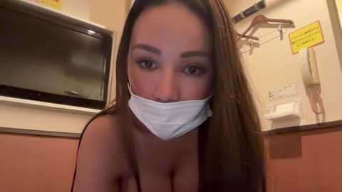 Media: Video of a woman with long brown hair and a white surgical mask, revealing her bare breasts, in a dimly lit room with a television and yellow sign.