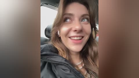 Media: A candid video of a young woman with light skin and wavy brown hair, smiling broadly, wearing a black jacket, taken inside a car.