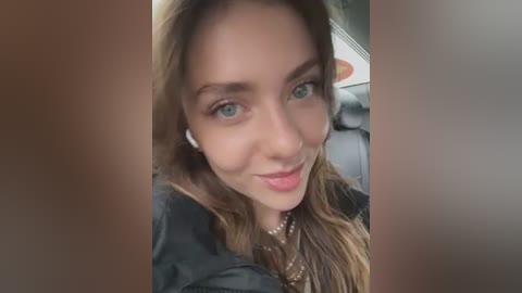 Media: Video of a young woman with light skin, brown hair, and green eyes, wearing a black jacket, smiling slightly, with blurred background of a car interior.