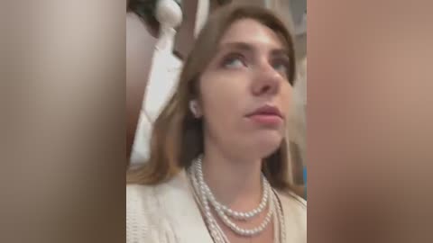 Media: A video of a young woman with fair skin, light brown hair, and blue eyes, wearing multiple pearl necklaces and a white cardigan, looking contemplative. The background is blurred.