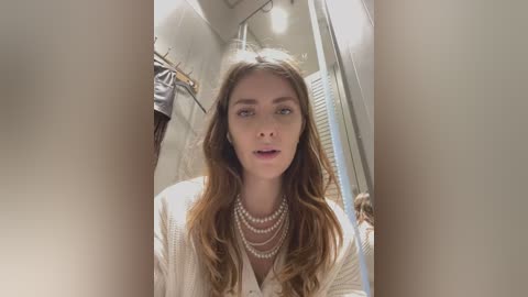 Media: Video of a young woman with long brown hair, fair skin, wearing a cream-colored blouse, pearls, and a casual expression. She is indoors in a cluttered, dimly lit bathroom with a mirror and hanging clothes.